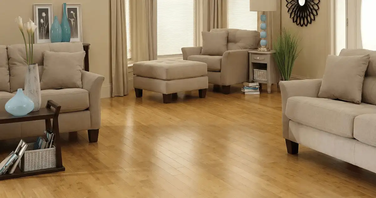 A living room with professionally installed bamboo flooring, beige sofas, a matching ottoman, a side table with a blue lamp, vases, framed pictures, and large windows with blinds and curtains in Fort Wayne, IN