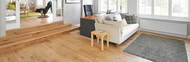 A modern living room with professionally installed laminate flooring, a beige sofa, a small side table, a gray rug, and large windows with white shutters in Fort Wayne, IN